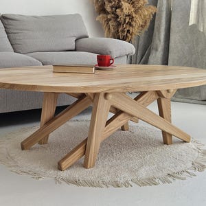 Könnte beinhalten: Ein ovaler Couchtisch aus hellem Holz mit einem einzigartigen, abgewinkelten Bein-Design. Eine rote Tasse und ein Buch liegen auf der Tischoberfläche. Der Tisch steht auf einem runden, gefransten Teppich in einem neutralen Wohnzimmer.