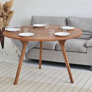 Pode incluir: Uma mesa de jantar redonda de madeira com acabamento natural, com pratos brancos, copos transparentes e talheres. A mesa tem tr&ecirc;s pernas anguladas. Um sof&aacute; cinza est&aacute; ao fundo. Um vaso decorativo com plantas secas est&aacute; &agrave; esquerda.