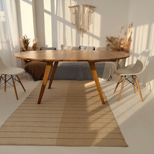 May include: A light wood oval dining table with four angled legs, set on a striped rug. Two white chairs with black and wood legs are on either side. The room has a neutral color palette with natural light.