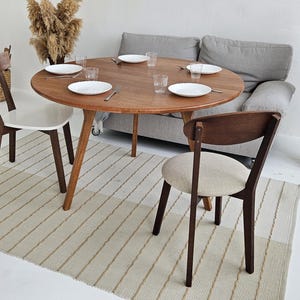 Pode incluir: Uma mesa de jantar redonda de madeira com seis pratos brancos, copos e talheres. Tr&ecirc;s cadeiras de madeira com assentos brancos e bege circundam a mesa. Um sof&aacute; cinza e um tapete listrado tamb&eacute;m est&atilde;o na sala.