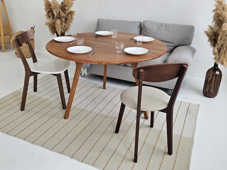 Pode incluir: Uma mesa de jantar redonda de madeira com seis pratos brancos, copos e talheres. Tr&ecirc;s cadeiras de madeira com assentos brancos e bege circundam a mesa. Um sof&aacute; cinza e um tapete listrado tamb&eacute;m est&atilde;o na sala.