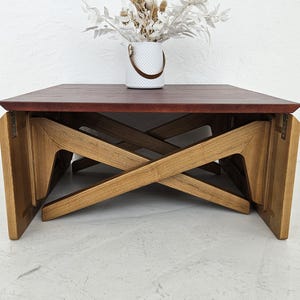 May include: A wooden coffee table with a burgundy top and a unique X-shaped base. The table is made of natural wood and has a rustic, vintage look.