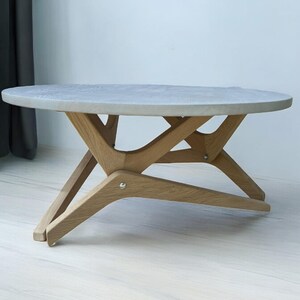 Può includere: Un tavolino da caffè rotondo in cemento grigio chiaro con una base in legno a forma di stella a raggiera. Il tavolo ha una finitura in legno naturale ed è sostenuto da quattro gambe.
