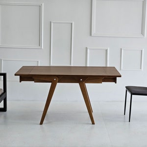 Puede incluir: Una mesa de madera marr&oacute;n con una extensi&oacute;n de hoja abatible y una silla de metal negro. La mesa tiene un dise&ntilde;o simple con cuatro patas y una tapa rectangular. La silla tiene un marco de metal negro y un asiento de tela gris.