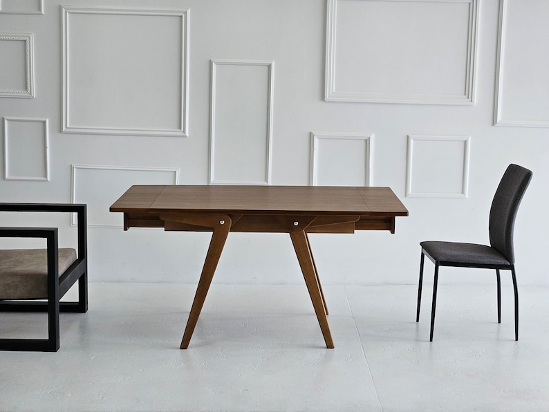 Puede incluir: Una mesa de madera marr&oacute;n con una extensi&oacute;n de hoja abatible y una silla de metal negro. La mesa tiene un dise&ntilde;o simple con cuatro patas y una tapa rectangular. La silla tiene un marco de metal negro y un asiento de tela gris.