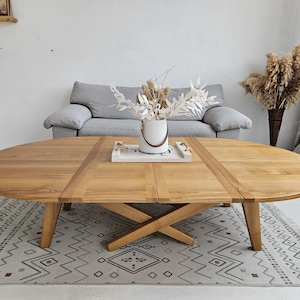 May include: Oval wooden coffee table with a natural finish. The table has a cross-shaped base and is set on a patterned rug. A gray sofa and decorative vase with dried flowers are in the background.