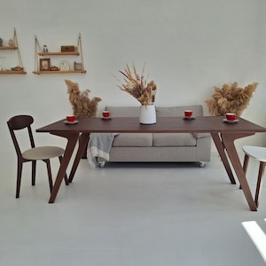 May include: A contemporary dining area with a dark brown rectangular table and angled legs, accompanied by two chairs. A grey sofa and a white vase with dried flowers are in the background. Red cups are placed on the table.