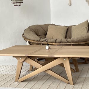 Könnte beinhalten: Ein heller Holz-Klapptisch mit einem Kreuzfuß. Der Tisch steht in einem Wohnzimmer mit einem großen, runden Korbstuhl mit Kissen.