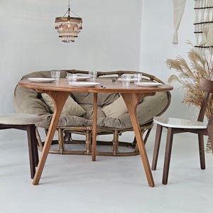 Pode incluir: Uma mesa de jantar de madeira com um design de pernas &uacute;nico, com pratos e copos. Duas cadeiras de madeira com assentos estofados e um sof&aacute; de rattan s&atilde;o dispostos ao redor da mesa. Uma lumin&aacute;ria pendurada acima, e elementos decorativos.