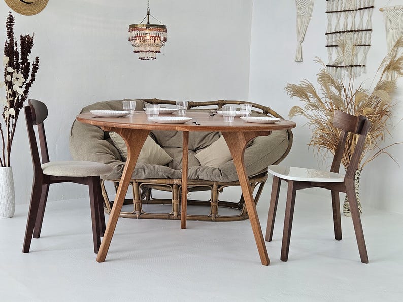 Pode incluir: Uma mesa de jantar de madeira com um design de pernas &uacute;nico, com pratos e copos. Duas cadeiras de madeira com assentos estofados e um sof&aacute; de rattan s&atilde;o dispostos ao redor da mesa. Uma lumin&aacute;ria pendurada acima, e elementos decorativos.