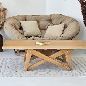 May include: A light brown wooden coffee table with a unique X-shaped base. The table has a rectangular top and is sitting on a white rug with a geometric pattern.