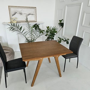 Puede incluir: Una mesa de comedor de madera con una tapa marr&oacute;n y cuatro patas. Dos sillas est&aacute;n colocadas en la mesa, una con un asiento negro y la otra con un asiento gris.
