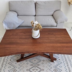 Könnte beinhalten: Ein Couchtisch aus massivem, dunkelbraunem Holz mit natürlicher Kante. Der Tisch steht auf einem gemusterten Teppich. Auf dem Tisch steht eine weiße Vase mit getrockneten Blumen. Der Tisch ist ca. 150 cm lang.
