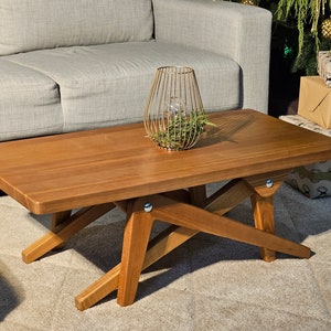 Op de afbeelding: Een houten salontafel met een rechthoekig blad en een unieke X-vormige basis. De tafel is gemaakt van lichtbruin hout en heeft een natuurlijke afwerking. Een gouden metalen kaarsenhouder met een glazen kaars staat op de tafel.