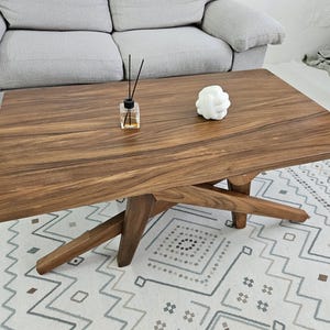 May include: A dark brown wooden coffee table with a unique X-shaped base. The table has a smooth, polished surface and is sitting on a white and grey patterned rug.