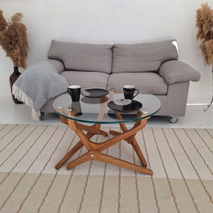 May include: A living room setting with a round glass coffee table with a wooden base. Two black mugs and plates are on the table. A grey sofa, a striped rug, and decorative plants complete the scene.