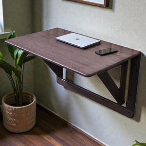 May include: A dark brown, wall-mounted folding desk with a laptop and smartphone on top. The desk has a triangular support structure. A potted plant and a window are visible in the background.