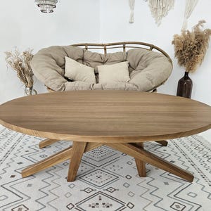 May include: A large, oval-shaped wooden coffee table with a natural finish. The table has a starburst-style base and sits on a patterned rug. The background includes a woven chair, pillows, and decorative elements, creating a cozy living room setting.