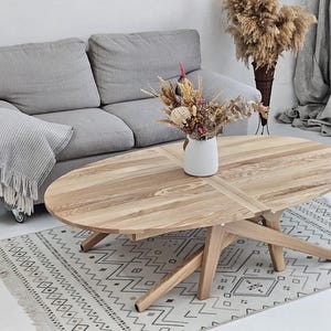 May include: An oval wooden coffee table with a natural finish, featuring a four-legged base and a decorative floral arrangement in a white vase. The table is approximately 150 cm long. A grey sofa and patterned rug complete the scene.