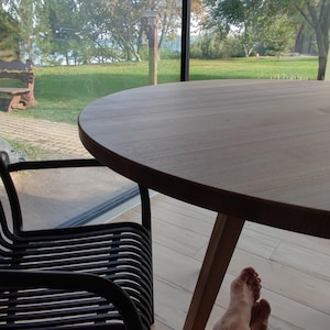 May include: A person sits at a wooden table with their legs crossed. The table is round and has a natural wood finish. The person is wearing blue shorts with a white pattern. The table is in front of a large window with a view of a green lawn and trees.