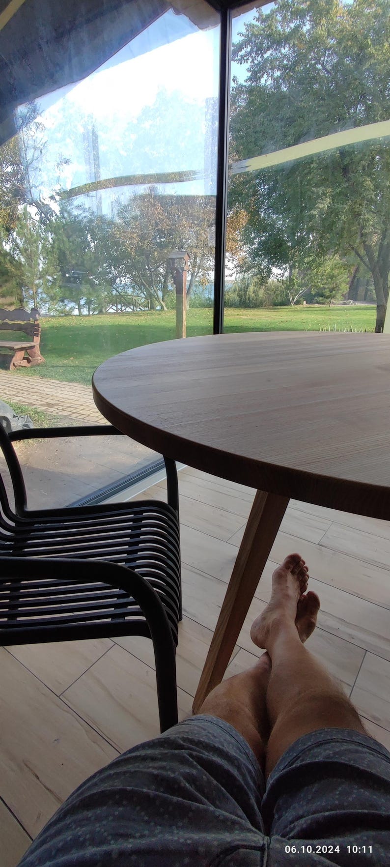 May include: A person sits at a wooden table with their legs crossed. The table is round and has a natural wood finish. The person is wearing blue shorts with a white pattern. The table is in front of a large window with a view of a green lawn and trees.
