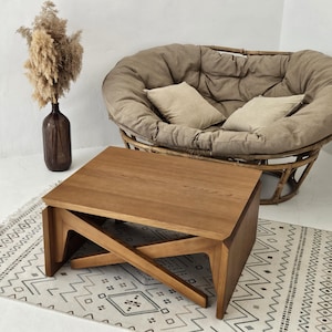 May include: A wooden coffee table with a distinctive X-shaped base. The table is crafted from natural wood and has a light brown finish. It is positioned on a white and black patterned rug.