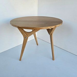 Puede incluir: Una mesa redonda de madera con un acabado marrón claro y una base de trípode única y angulada.