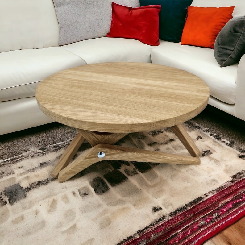 May include: A light wood round coffee table with a unique, foldable design. The table has a natural wood finish and is sitting on a patterned rug.