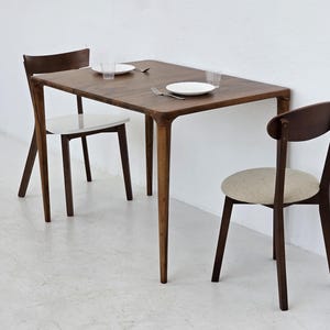 May include: A rectangular dark wood dining table with four tapered legs and two matching chairs. One chair features a white seat, while the other has a beige upholstered seat. The table is set with two white plates, two clear glasses, and two forks.