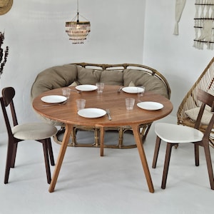 Pode incluir: Uma mesa de jantar redonda de madeira com quatro cadeiras, pronta para uma refei&ccedil;&atilde;o. A mesa &eacute; rodeada por um sof&aacute; de vime e uma cadeira de vime. A sala &eacute; decorada em tons neutros com uma lumin&aacute;ria suspensa.