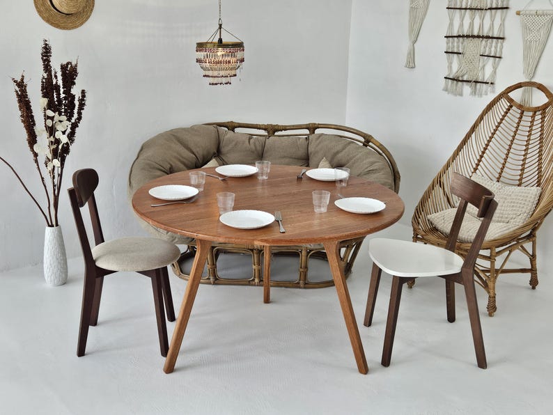 Pode incluir: Uma mesa de jantar redonda de madeira com quatro cadeiras, pronta para uma refei&ccedil;&atilde;o. A mesa &eacute; rodeada por um sof&aacute; de vime e uma cadeira de vime. A sala &eacute; decorada em tons neutros com uma lumin&aacute;ria suspensa.