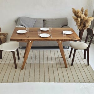 May include: A wooden dining table with a natural edge, set with white plates and glasses. Two chairs with white seats and two with brown seats surround the table. A beige rug and a grey sofa are in the background. A vase of dried flowers is on the right.