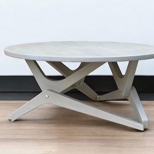 May include: A light gray round coffee table with a unique, geometric, X-shaped base. The table is made of wood and has a smooth, polished finish.