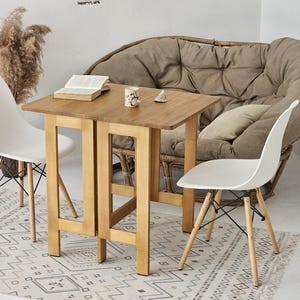 May include: A light wood folding table with two white chairs. The table is set with a book and a decorative object. The chairs have wooden legs and are placed on a patterned rug.