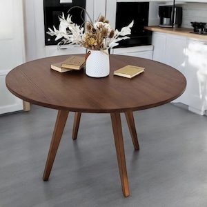 Op de afbeelding: Een ronde houten tafel met drie poten. De tafel is bruin en heeft een glad oppervlak. Er staat een vaas met gedroogde bloemen op de tafel, samen met twee boeken.
