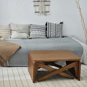 May include: A wooden coffee table with a unique X-shaped base. The table is brown and has a rectangular top. It is sitting on a white and beige striped rug in front of a couch with black and white patterned pillows.
