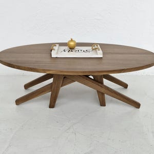 May include: A wooden oval coffee table with a natural brown finish. The table has a unique X-shaped base and features a white tray with the word "Home" written on it.