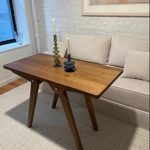 May include: A wooden table with a light brown finish and a simple design. The table has three legs on each side and a rectangular top. Two candle holders with candles are on the table. One candle holder is green glass and the other is blue glass.