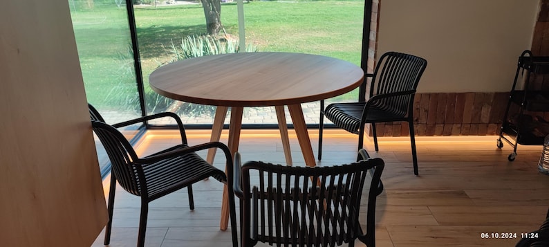 May include: A round wooden table with four black chairs. The table and chairs are in a room with a large window overlooking a grassy area.