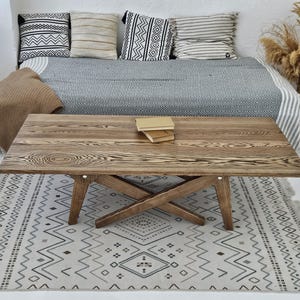 May include: A wooden coffee table with a natural wood grain finish. The table has a cross-legged base and sits on a patterned rug. Pillows and a blanket are on a bed in the background. Two books are on the table. The table is approximately 120 cm long.