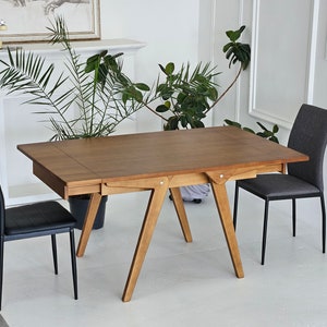 May include: A wooden dining table with a brown finish and a leaf extension. The table has a simple, modern design with a rectangular top and four legs. Two black chairs and one gray chair are placed around the table.