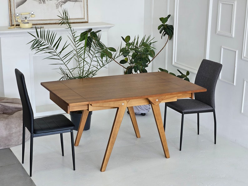 Puede incluir: Una mesa de comedor de madera con un acabado marr&oacute;n y una extensi&oacute;n de hoja. La mesa tiene un dise&ntilde;o simple y moderno con una tapa rectangular y cuatro patas. Dos sillas negras y una silla gris se colocan alrededor de la mesa.