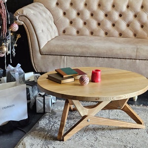 May include: A round wooden coffee table with a natural wood finish and a unique X-shaped base. The table is decorated with two red candles and a stack of books.