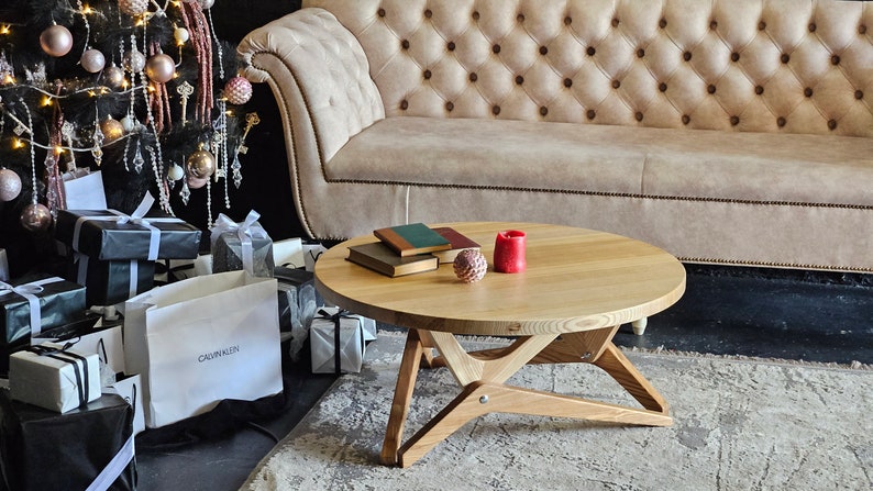 May include: A round wooden coffee table with a natural wood finish and a unique X-shaped base. The table is decorated with two red candles and a stack of books.