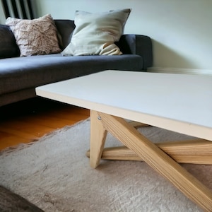 Könnte beinhalten: Ein weißer Couchtisch mit einem Untergestell aus hellem Holz. Das Untergestell besteht aus mehreren Holzstücken, die in einem Kreuzmuster zusammengefügt sind.