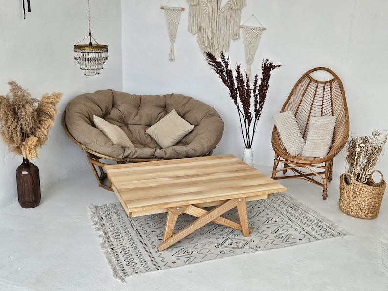 May include: A light wood coffee table with a unique X-shaped base. The table is set on a white and grey patterned rug in a room with a brown and beige beanbag chair and a wicker chair with white cushions.