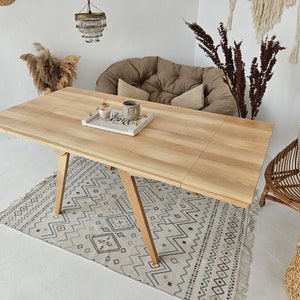 May include: A light wood dining table with a natural finish and a geometric pattern rug underneath. The table has a rectangular top and four legs with a triangular shape. There is a small tray on the table with two candles and a ceramic mug.