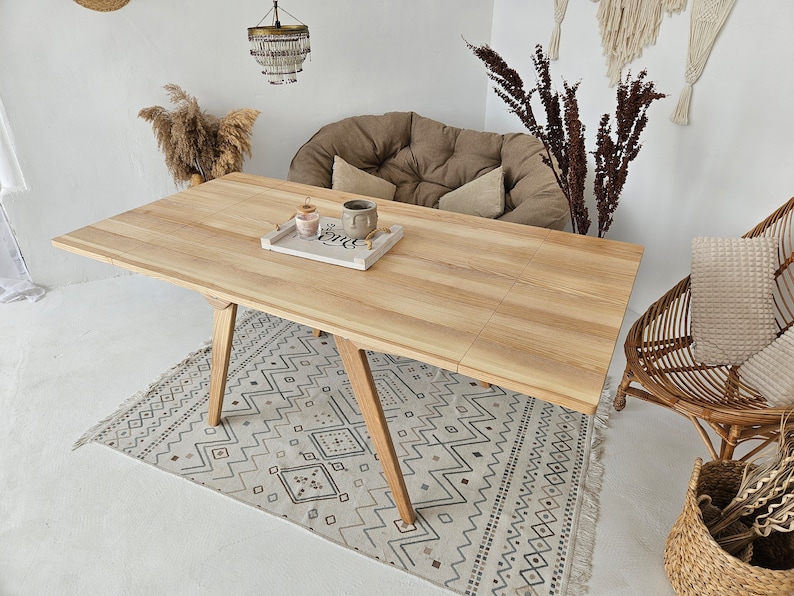 May include: A light wood dining table with a natural finish and a geometric pattern rug underneath. The table has a rectangular top and four legs with a triangular shape. There is a small tray on the table with two candles and a ceramic mug.