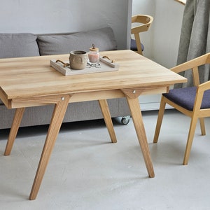 May include: A light wood table with a rectangular top and angled legs. The table has a small tray on top with a ceramic mug and a glass jar. A grey sofa is behind the table. Two light wood chairs with dark grey cushions are in the foreground.