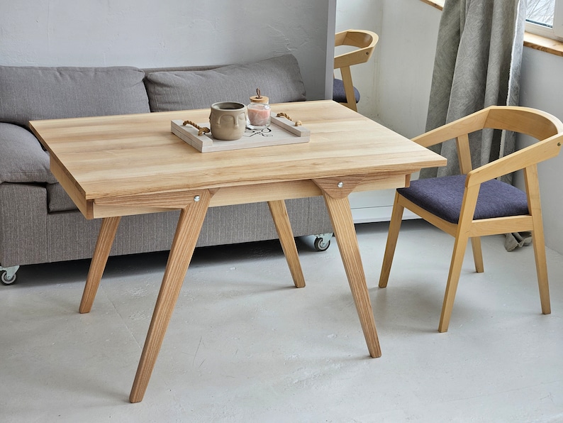 May include: A light wood table with a rectangular top and angled legs. The table has a small tray on top with a ceramic mug and a glass jar. A grey sofa is behind the table. Two light wood chairs with dark grey cushions are in the foreground.